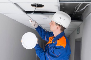 Mavi üniformalı bir ustabaşı, sahte tavanların ışıklarını yakarken sorun çıkarıyor, lamba bağlantı kutusunun bağlantılarını kontrol ediyor.