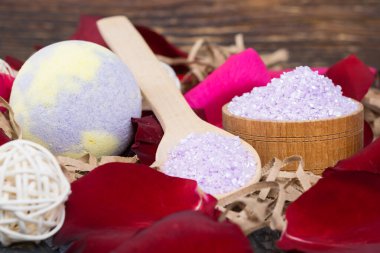 Koyu renk ahşap bir arka planda, gül yapraklarıyla çevrili, bir kabın içinde ve bir kaşıkta, mis kokulu banyo tuzu ve gevşetici bir bomba.