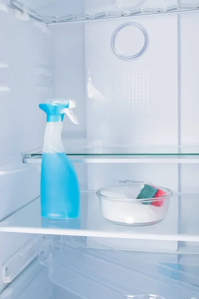 against the background of a white refrigerator, on a shelf, there is a container with cleaning foam, sponge and spray in a bottle