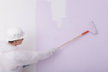 Koruyucu giysili ve kasklı bir müteahhit, açık mor renkli bir silindirle duvarı boyuyor.