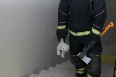 Bir itfaiyeci elinde oyuncak bir oyuncak ve kapı açmak için baltayla merdivenleri tırmanır.