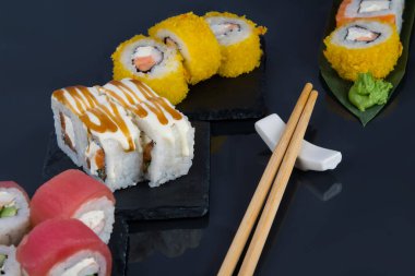 Yakın plan, siyah bir masanın üzerinde çeşitli Japon dürümleri, ton balığı, yumuşak peynir ve teriyaki sosu ve tempura m ve ahşap çubuklarda fırınlanmış rulo var.