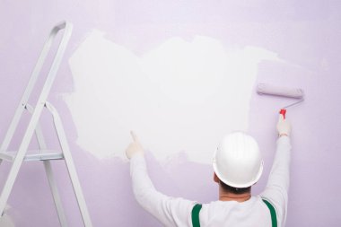 Sert şapkalı bir işçi beyaz bir duvar eflatunu boyuyor.