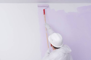 Koruyucu giysili ve kasklı bir inşaat işçisi, açık mor renkli, dikiz görüşlü beyaz bir duvar boyuyor.