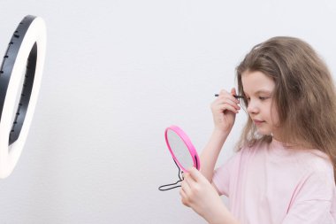 Blogcu bir kız, dairesel bir ışığın önünde makyaj yapar, göz kapaklarına gölgeler koyar, pembe bir aynaya bakar.