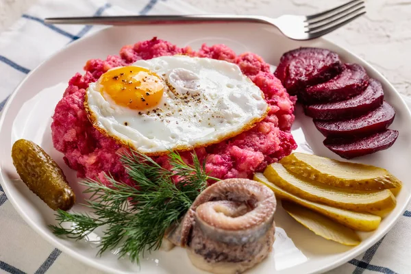 labskaus, corned beef, mashed potatoes with the beetroot topped with fried eggs, sliced beets, with pickles, and rollmops, pickled, rolled herring on a plate, german cuisine