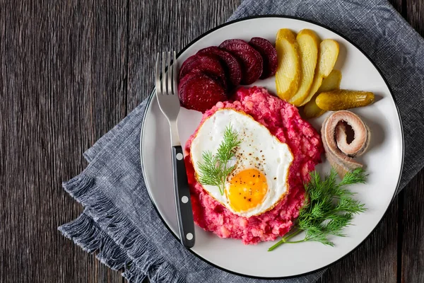 labskaus, corned beef, mashed potatoes with the beetroot topped with fried eggs, sliced beets, with pickles, and rolled herring on a plate, german cuisine, hamburg style,  flat lay