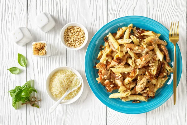 chicken alfredo pasta penne with caramelized onion sprinkled with pine nuts and grated parmesan cheese on a blue plate on white wooden table, horizontal view from above, flat lay