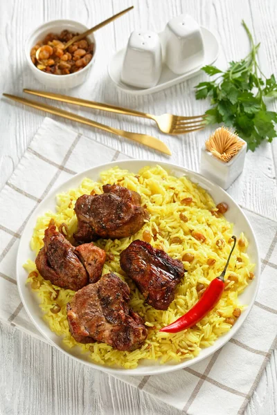 Mutton Zafrani Pulao, roasted lamb meat served with raisins saffron rice on a white plate on a white wooden table, vertical view from above