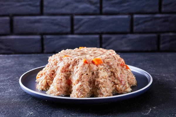 pork and chicken meat aspic, jellied slow cooked meat on a plate on a concrete table with a black brick wall at the background, close-up