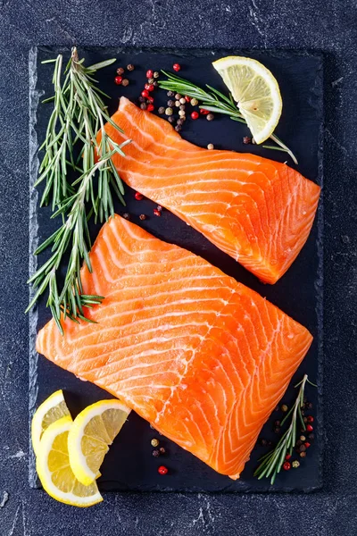 salmon fillet pieces with peppercorn, rosemary and lemon on black slate plate on a grey concrete table, vertical view from above, flat lay