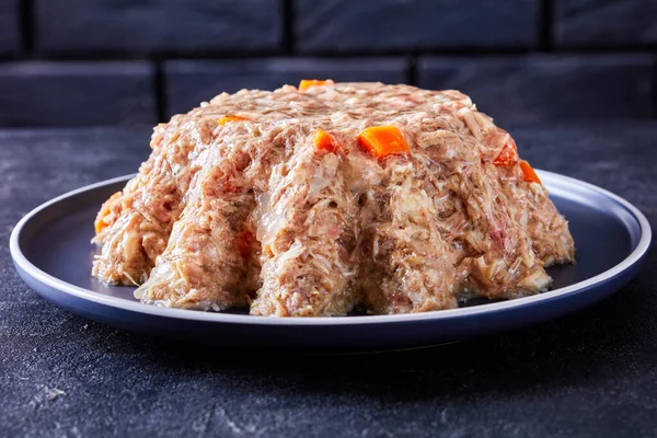 pork and chicken meat aspic, jellied slow cooked meat on a plate on a concrete table with a black brick wall at the background, close-up