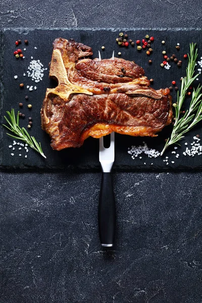 fried porterhouse steak, t-bone steak is speared with carving fork on black plate with thyme, rosemary and peppercorn on concrete table, vertical view from above, flat lay, free space