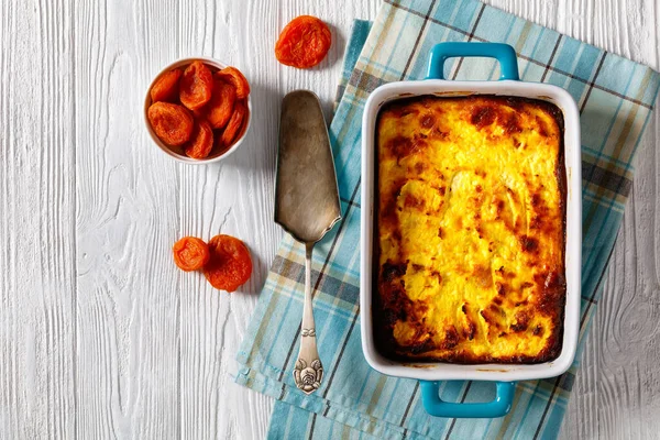 Cottage Cheese Breakfast Bake with dried apricot in a baking dish on a white wooden table with ingredients, flat lay, free space