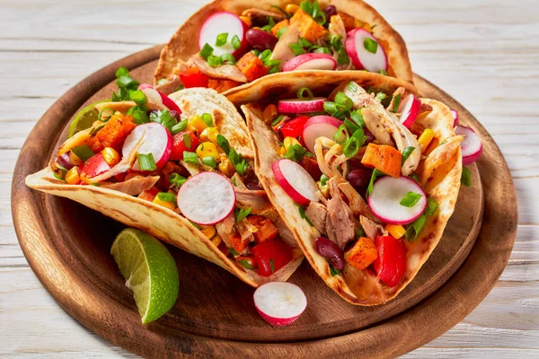 close-up of pitacos, mexican tacos with pita bread, spicy pulled chicken meat, red kidney beans, corn, radish, red pepper, roast sweet potatoes and lime on wooden board