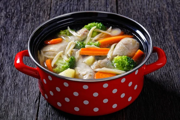 chicken vegetable soup with broccoli, carrots, parsnip, leek and pasta in red pot on dark wooden table, horizontal view from above