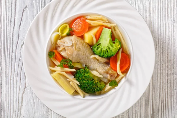 chicken spring soup with broccoli, carrots, parsnip, leek and pasta in white bowl on white wooden table, horizontal view from above, flat lay, close-up