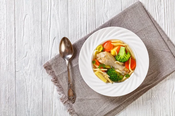 chicken spring soup with broccoli, carrots, parsnip, leek and pasta in white bowl on white wooden table, horizontal view from above, flat lay, free space