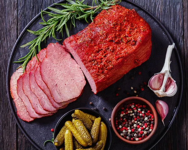 fresh sliced beef pastrami  sliced on black plate with fresh rosemary,garlic and pickles on dark table, horizontal view from above, flat lay, close-up