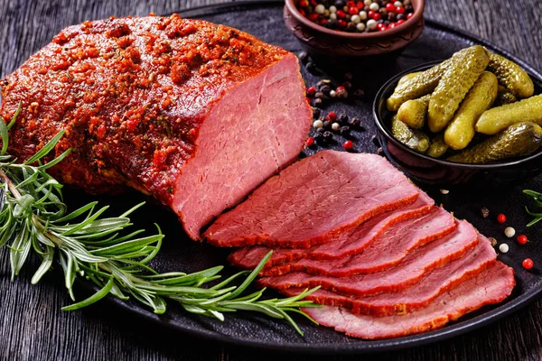 fresh sliced beef pastrami  sliced on black plate with fresh rosemary, thymes and pickles on dark table, horizontal view from above, close-up