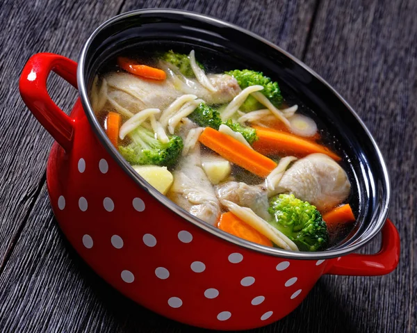 chicken vegetable soup with broccoli, carrots, parsnip, leek and and pasta in red pot on dark wooden table, horizontal view from above, close-up