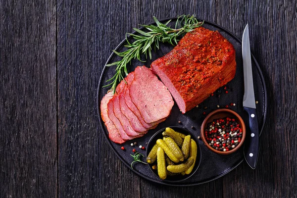 fresh sliced beef pastrami  sliced on black plate with fresh rosemary, thymes and pickles on dark table, horizontal view from above, flat lay, free space