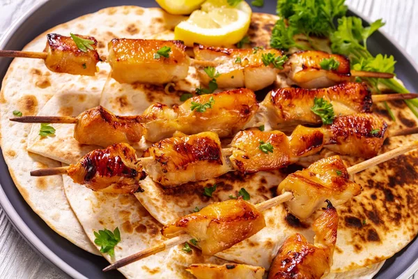 grilled chicken skewers sprinkled with chopped fresh parsley over flatbreads on plate on white wooden table, close-up