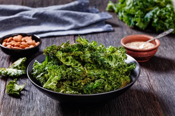 kale chips in black bowl with almonds, sesame seeds, cloth on dark oak textured table, horizontal view from above
