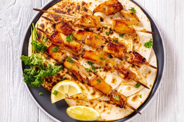 bbq chicken kebabs sprinkled with chopped fresh parsley over flatbreads on plate on white wooden table, flat lay, close-up, macro
