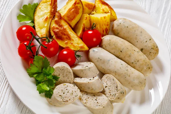 close-up of Weisswurst, bavarian white sausage of minced veal, pork back bacon, spices and parsley on white plate with roast potato, fresh tomatoes, horizontal view