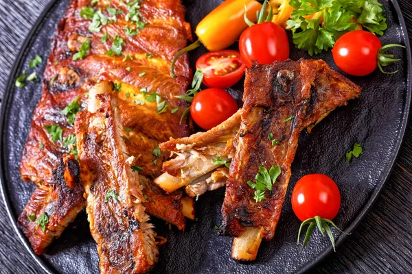 Fall Off the Bone Oven Baked Ribs with fresh tomatoes, chili peppers on black platter on dark wooden table, horizontal view from above, close-up