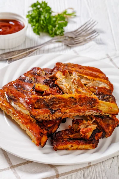 Fall Off the Bone Oven Baked Ribs on white plate with ketchup on white wooden table, vertical view from above, close-up
