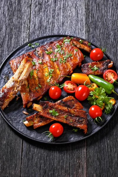 Oven Baked Ribs with fresh tomatoes, chili peppers on black platter on dark wooden table, vertical view from above