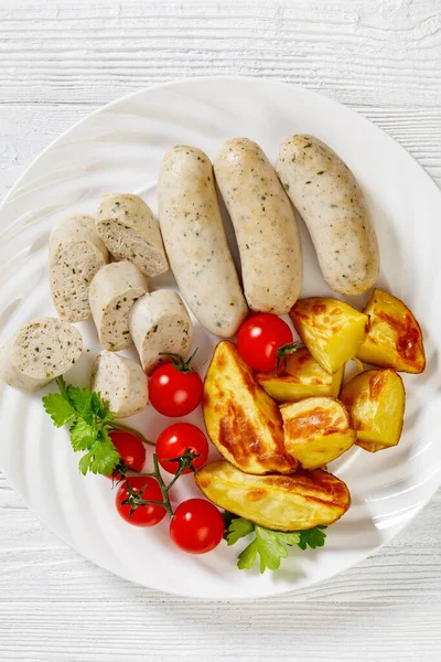 Weisswurst, bavarian white sausage of minced veal, pork back bacon, spices and parsley on white plate with roast potatoes, fresh tomatoes, vertical view from above, close-up