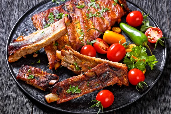 Fall Off the Bone Oven Baked Ribs with fresh tomatoes, chili peppers on black platter on dark wooden table, horizontal view from above