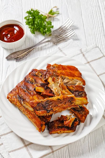 Fall Off the Bone Oven Baked Ribs on white plate with ketchup on white wooden table, vertical view from above