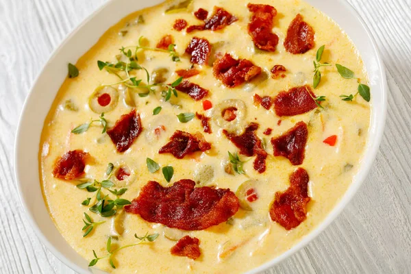 close-up of Beer cheese soup with red pepper stuffed green olives, vegetables and fried bacon in white bowl on white wooden table, american cuisine