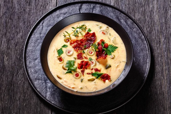 Beer cheese soup with red pepper stuffed green olives, vegetables and fried bacon in black bowl on dark wooden table, landscape view