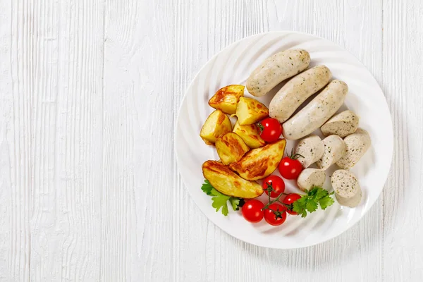 Weisswurst, bavarian white sausage of minced veal, pork back bacon, spices and parsley on white plate with roast potatoes, fresh tomatoes, horizontal view from above, flat lay, free space