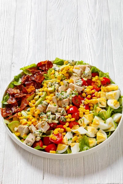American garden salad of chopped romaine lettuce, tomato, crisp bacon, chicken breast, hard-boiled eggs, avocado, blue cheese, and red-wine vinaigrette on plate, vertical view, close-up