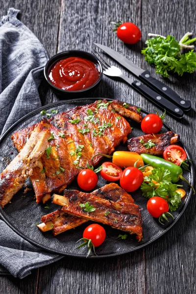Fall Off the Bone Oven Baked Ribs with fresh tomatoes, chili peppers on black platter on dark wooden table, vertical view from above
