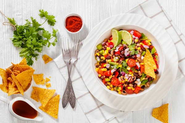 Black Bean Salad with Black-Eyed Peas, pepper, red onions, corn, cherry tomatoes and cilantro in white bowl with tortilla chips, nachos and vinaigrette dressing on wood table, flat lay