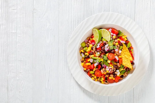 Black Bean Salad with Black-Eyed Peas, pepper, red onions, corn, cherry tomatoes and cilantro in white bowl with tortilla chips, nachos  on wood table, flat lay, free space