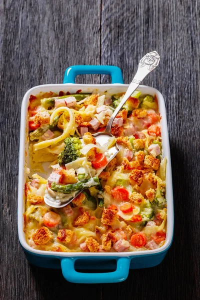 Ham and Noodle Casserole with egg noodles, chopped ham, carrots, broccoli, asparagus and a rich and creamy sauce, topped with croutons in baking dish with cake shovel, vertical view