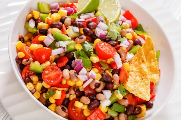 close-up of Black Bean Salad with Black-Eyed Peas, pepper, red onions, corn, cherry tomatoes and cilantro in white bowl with tortilla chips, nachos
