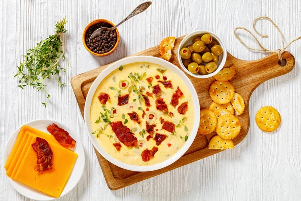 Beer cheese soup with red pepper stuffed green olives, vegetables and fried bacon in white bowl on chopping board with salt crackers on white wooden table, flat lay