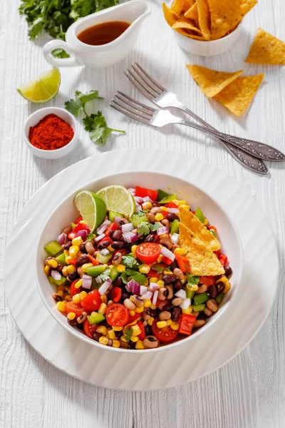 Black Bean Salad with Black-Eyed Peas, pepper, red onions, corn, cherry tomatoes and cilantro in white bowl with tortilla chips, nachos, vertical view, close-up