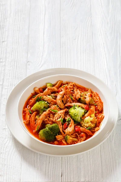 slow-cooked shredded chicken breast stew with tomato sauce, red pepper, onion and broccoli in white bowl on wood table,vertical view, copy space