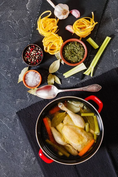 cooked chicken bouillon of whole chicken, celery, onion and carrot in red pot with ingredients and tagliatelle pasta on concrete table, vertical view from above