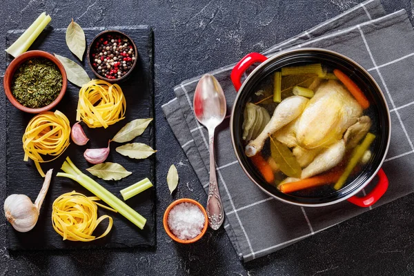 hot chicken bouillon of whole chicken, celery, onion and carrot in red pot with ingredients and tagliatelle pasta on concrete table, landscape view from above, flat lay